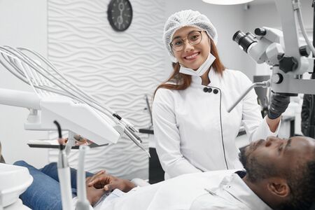 Beautiful female dentist looking at camera, smiling and posing while curing teeth of patient. Young woman working while african man lying on dentist chair in clinic. Concept of health and treatment.の写真素材