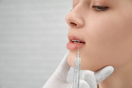 Closeup of beautiful lady posing while female cosmetologist doing beauty injections in clinic. Young woman looking down while doctor plumping lower lip. Concept of collagen and cosmetics.の写真素材