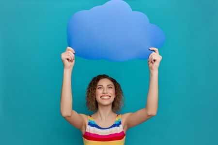 Young, positive, happy girl with curly hair holding blue paper speech bubble in form of cloud above...