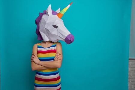 Young, creative girl holding on head 3D white unicorn head, posing on blue background in studio. Slender, stylish model standing in striped dress holding crossed hands.の写真素材
