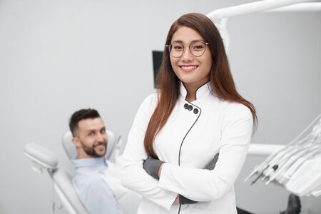 Front view of beautiful female dentist in white uniform looking at camera and posing while male patient lying on dentist chair at background. Professional doctor working in clinic.の写真素材