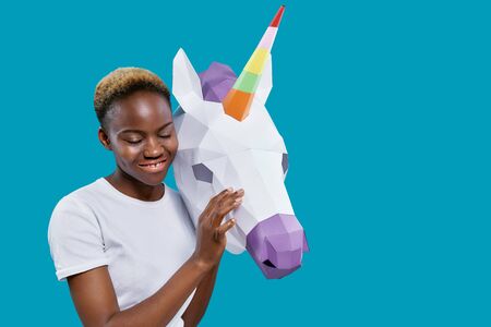 Cheerful african woman holding paper unicorn head on blue background with copy space. Positive girl in white t shirt posing with white 3D unicorn, looking down, smiling.の写真素材