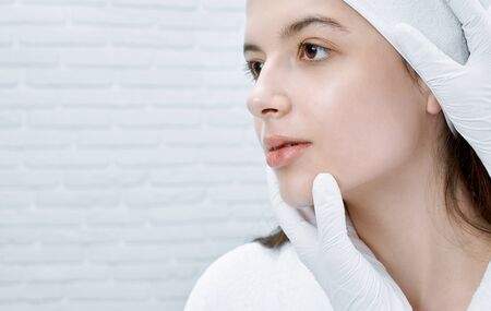 Young, beautiful woman in white bathrobe receiving procedure of inspecting face in cosmetology center. Hands of doctor in white medical gloves touching chin. Concept of skin care.の写真素材