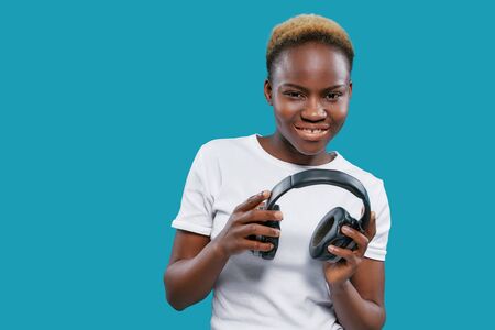 Beautiful, cheerful, charming african woman with short hair holding headphones. Pretty, positive, stylish model posing on blue background in studio, looking at camera.の写真素材