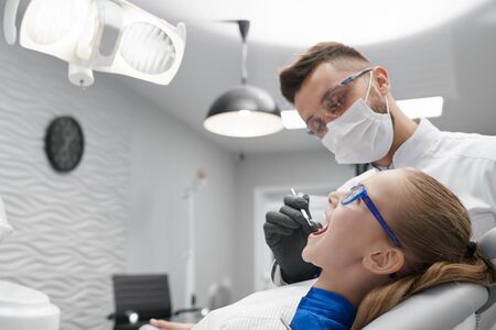 Side view of serious male dentist in mask and glasses brushing teeth of little patient in clinic. Pretty girl with open mouth lying on dentist chair. Concept of treatment and health.の写真素材