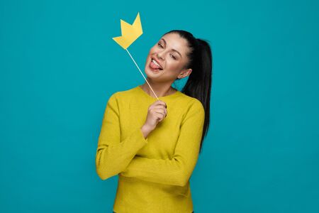 Funny young girl in yellow sweater keeping paper crown and showing tongue on blue isolated background. Excited woman looking at camera and posing in studio. Concept of happiness and fun.の写真素材