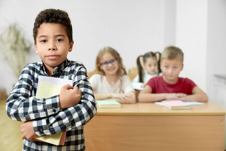 Handsome schoolboy standing in classroom, holding in hands books, looking at camera, posing. Cheerful pupils sitting at desk on background.の写真素材