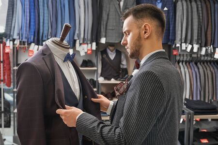 Young, handsome man choosing suit in fashionable boutique. Jackets of fashionable prints on hangers in row, clothing and footwear on shelves. Male clothing on mannequin.の写真素材