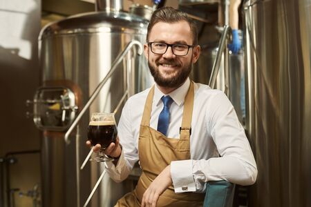 Positive, cheerful brewer sitting on steps of metal platform, holding glass with dark beer. Handsome worker smiling and posing on background of steel storage tank.の写真素材