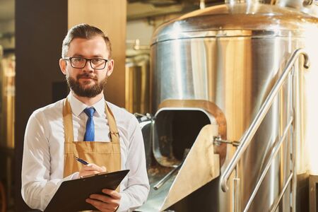 Adult expert monitoring brewery process and writing. Handsome, bearded man in glasses, white shirt, tie and brown apron holding folder and pen, posing, looking at camera.の写真素材