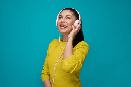 Happy, cheerful model in yellow jumper listening music in white headphones. Pretty, beautiful,...