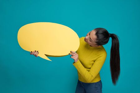 Beautiful, stylish, charming model holding yellow paper speech bubble. Pretty, gorgeous, young woman in yellow jumper and blue jeans looking at speech bubble.の写真素材