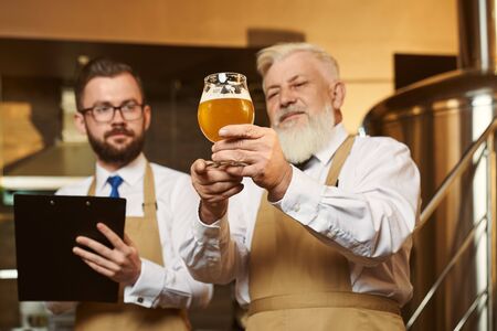 Two professional men wearing white shirts and aprons keeping glass of light beer and examining quality of alcohol. Colleagues looking at drink and writing in folder. Concept of brewery.の写真素材