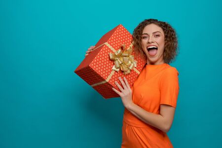 Positive young woman in orange dress looking at camera and shouting while keeping big present in hands. Happy female posing and laughing on blue isolated background. Concept of surprise.の写真素材