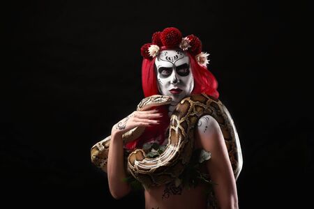 Sugar skull model posing with big snake around neck. Girl with red hair, red eyes and lips, red and...