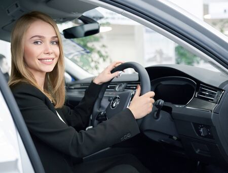 Young woman sitting in right handed automobile, looking at camera and smiling in auto showroom....