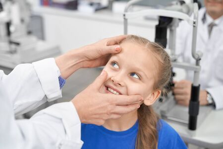 View from side of nice female oculist checking eye pupil of smiling girl in laboratory. Cheerful...