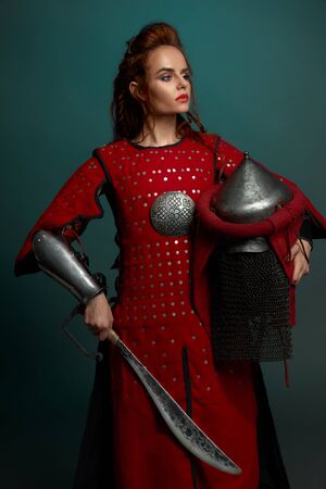 View form side of barbarian female fighter posing with helmet and knife in hands in studio. Young warrior at red armor looking aside on isolated background. Concept of confidence and weapon.の写真素材