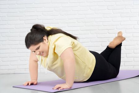 Brunette fat young girl in yellow hoodie and black leggings is trying to do push ups while standing...