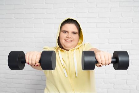 Smiling brunette chunky young lady in yellow hoodie looking at camera and holding two big black...