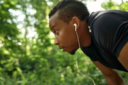 Close up of muscular runner with curly hair wearing in black t shirt outdoors. Handsome african sportsman listening music with headphones. Athlete posing side view, looking forward.の写真素材