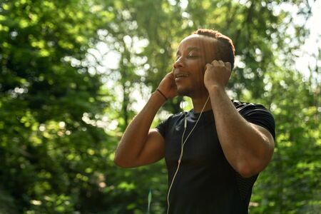 Close up of african sportsman listening music with headphones, enjoying. Muscular runner running in forest with closed eyes. Man with sportive body wearing in black t shirt.の写真素材