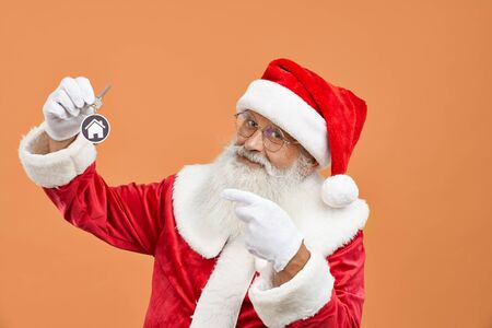 Portrait of happy senior Santa Claus with real grey beard and eyeglasses pointing on keys from new house or apartment on orange background. Christmas character in red costume giving present.の写真素材