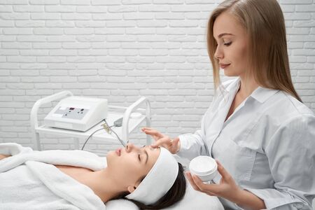 Beautiful doctor cosmetologist in white coat standing behind patient and applying cream on face....
