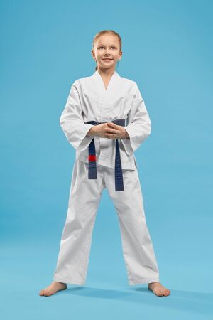Positive, cheerful girl standing in stance of karate on light blue background, looking up. Portrait of pretty junior wearing in white kimono with blue belt in studio. Concept of health and sport.の写真素材