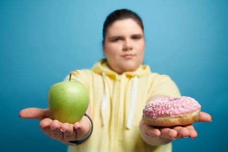 Close up of green fresh apple and donut with pink glaze which is holding young chunky brunette girl in yellow sweater. Concept of right choice of food in human life.の写真素材