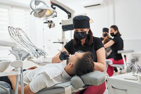 Side view of professional female doctor in uniform keeping probe and mirror and examining teeth of male patient. Man lying on chair with opened mouth in dental office. Concept of treatment and health.の写真素材