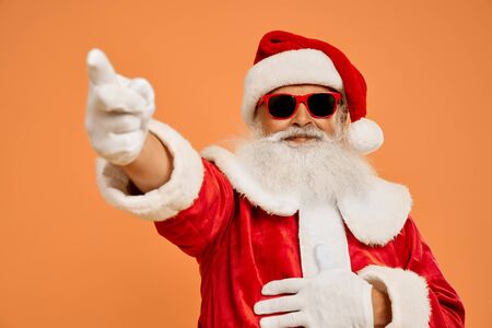 Senior bearded man in red Santa costume and trendy sunglasses over isolated background looking at camera and pointing on by finger. Front gesture and positive emotion.の写真素材