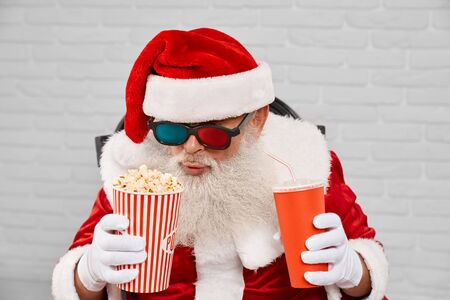 Senior man wearing traditional santa claus costume and 3d glasses, sitting in cozy armchair with popcorn and cola in white studio. Funny christmas character enjoying free time.の写真素材