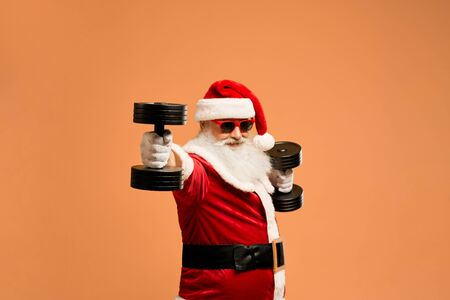 Optimistic elder modern Saint Nicholas in red winter costume, sunglasses and white gloves exercising with two black dumbbells, isolated on orange background. Senior man preparing for Christmasの写真素材