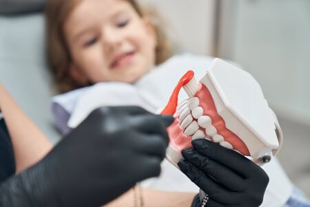 Dentist teaching girl how to brush teeth. Close up of dentist hands in black rubber gloves holding toothbrush and bruching teeth. Concept of dental care.の写真素材