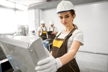 Selective focus of beautiful woman in protective mask and uniform looking at camera and posing while working with computer. Young engineer controlling equipment and devices. Concept of metalwork.の写真素材