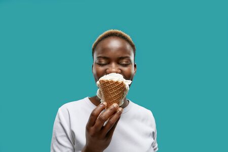 Front view of pretty african with closed eyes keeping waffle of ice cream and posing on blue...