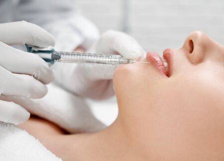 Close up of hands of cosmetologist in white rubber gloves making botox injection in female lips, holding syringe. Young beautiful woman receiving procedure for correction and beauty.の写真素材