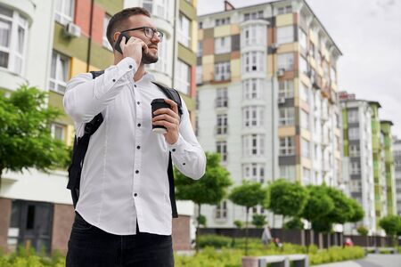 Close up of young bearded man talking by phone, holding plastic coffee cup on background of sleeping area in city. Student with backpack behind shoulder looking at multistory houses.の写真素材