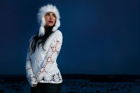 Fashionable model wearing in stylish warm hat and white knitted blouse posing on field on background...
