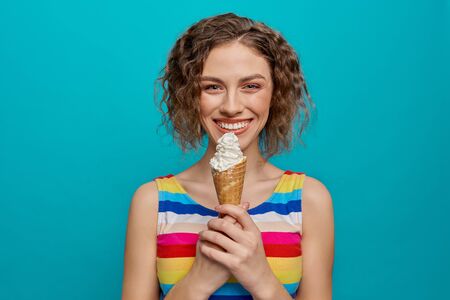 Happy, charming girl in striped dress holding ice cream wafer cone. Beautiful, cheerful, young woman with curly hair posing on blue background in studio, looking at camera, smiling.の写真素材