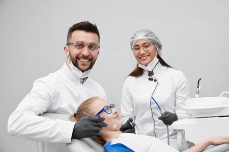 Happy dentists looking at camera and laughing while curing teeth of little client in clinic. Experienced male doctor brushing teeth and pretty female assistant helping. Concept of stomatology.の写真素材
