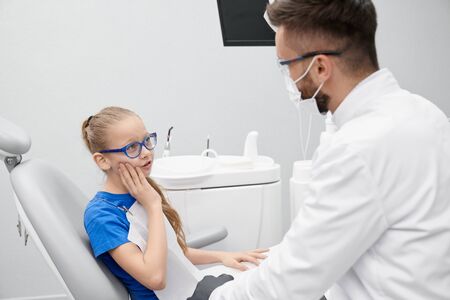 Side view of little patient complaining about pain in teeth in clinic. Girl in glasses sitting in dentist office, looking at doctor and talking. Concept of treatment and stomatology.の写真素材