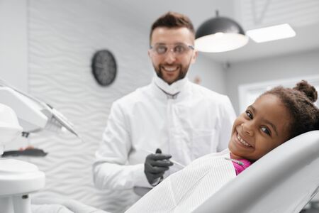 View from side of cheerful african child lying on dentist chair, looking at camera and smiling while doctor curing teeth. Professional dentist keeping tool and posing at background. Concept of care.の写真素材