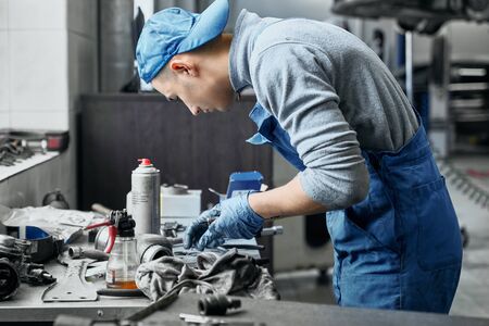 Side view of concentrated young car service employee working near desk with a lot of instruments on it. Repairman doing restoration of old details for disassembled carの写真素材