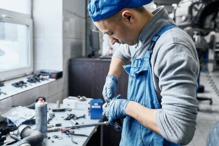 Repairman in car service wearing blue uniform and protective rubber gloves working near table with a lot of tools on it,preparing details to replace them in disassembled auto.Working process conceptの写真素材