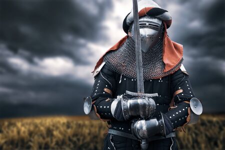 Front view of medieval knight in iron armor and headband posing and holding big sword. Portrait of...