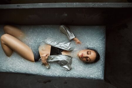 From above view of wet brunette girl lying in bath with muddy white water. Portrait of young female with perfect makeup in underwear and silver jacket posing with closed eyes. Concept of modeling.の写真素材