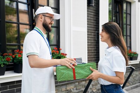 Side view of young bearded deliveryboy in white cap giving two gift boxes to woman. Pretty smiling brunette female in casual outfit receiving order and folder to sign near house. Delivery concept.の写真素材