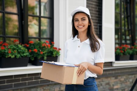 Front view of pretty happy joyful young female courier in white cap and casual outfit carrying box and folder. Brunette woman standing outdoors, holding parcel and smiling. Delivery concept.の写真素材